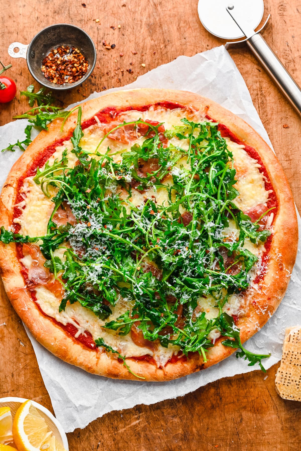 An arugula prosciutto pizza next to a dish of lemons and a pizza cutter. 