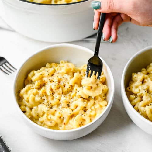 A black fork taking a bite of gouda mac and cheese in a white bowl.