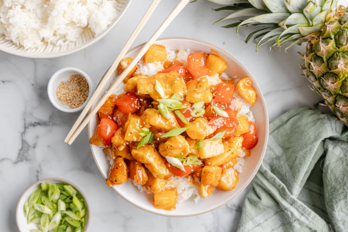 A bowl of sweet and sour chicken next to a dish of white rice and sliced green onions.